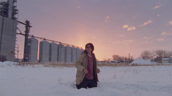 Een jonge man wadend door hoge sneeuw, met op de achtergond een olieraffinaderij tegen een zonsondergang.